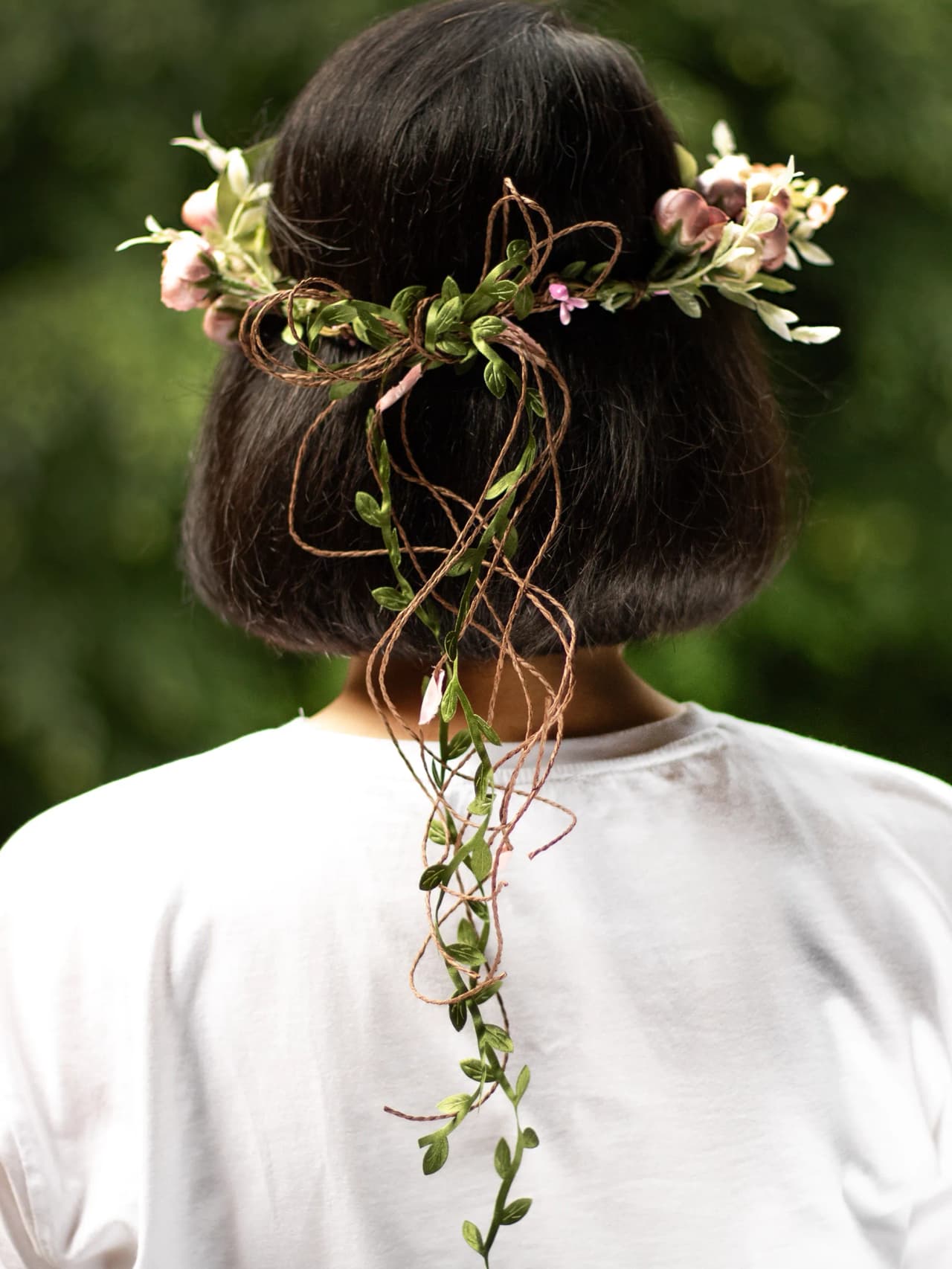 Person fotograferad bakifrån med en blomkrans i håret, dekorerad med rosa blommor, gröna blad och långa hängande snören av naturfärgad jute.