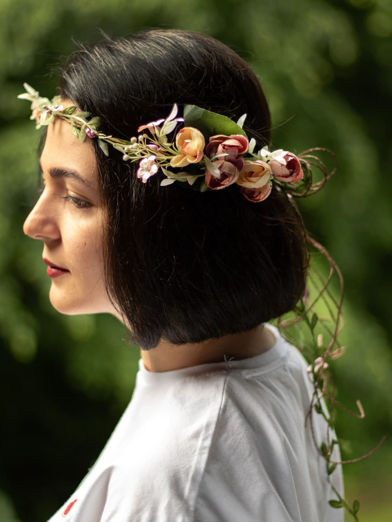 Sidovy av en blomkrans med rosa rosor, ljusa blad och slingrande jutesnören, placerad runt kort mörkt hår.